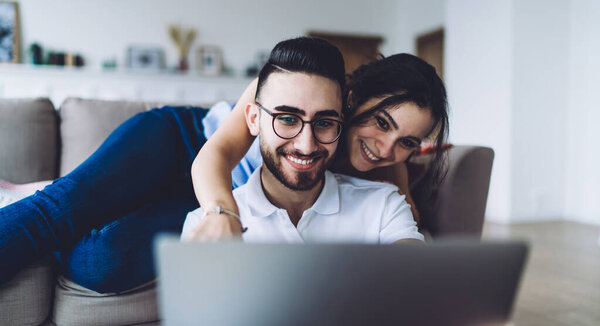 Young bearded smiling ethnic man sitting on floor leaning on sofa holding and browsing laptop while cheerful woman resting on sofa behind male and showing on screen at home