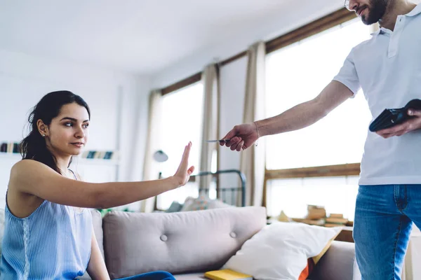 Smart confident woman gesturing with stretched hand sitting on sofa and refusing credit card suggesting by crop man standing in living room