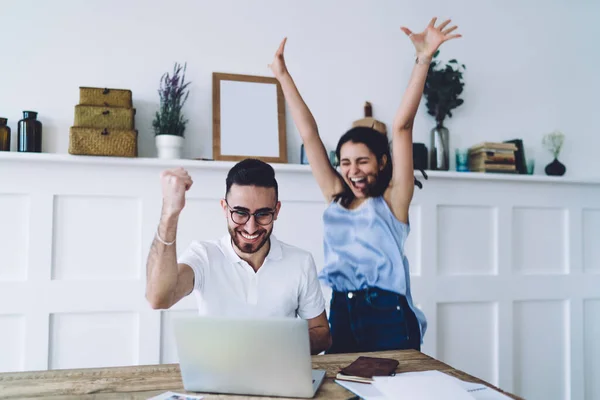 Young female in blue shirt and jeans jumping for joy with arms up and bearded man with toothy smile raising hand up with sign of success and looking at computer screen