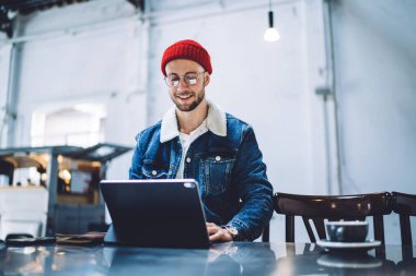 İnternette sörf yapmak ve bilgi aramak için dijital cihaz kullanan sıradan giyinmiş mutlu yakışıklı genç adam telefon ve bardakla çatı katında otururken...