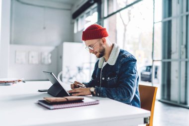 Yandan bakan genç erkek serbest grafik tasarımcı, kalın kıyafetler giyiyor, gözlük takıyor ve parlak beyaz odada tablet üzerinde çalışıyor.