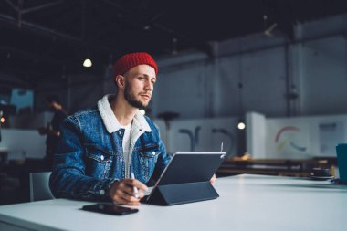 Yoğunlaşmış, şık, sakallı, erkek serbest çalışan, ofiste çalışan, kalın paltolu ve şapkalı, bulanık arkaplanlı tabletli bir adam. 