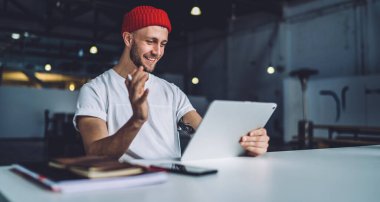 Kırmızı bereli genç adam oturmuş tabletle sohbet ediyor. Geniş endüstri ofisindeki masada çalışırken video görüşmesi yapıyor.