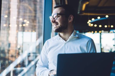 Mutlu gülümseyen iş adamı gözlük takıyor ve bilgisayarını kullanıyor. Ofisin geniş penceresinin yanında durup camdan şehri seyrediyor.