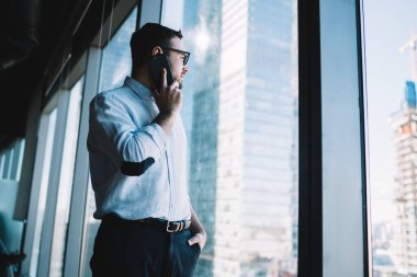 Profesyonel bir çalışanın resmi kıyafetleri içinde ofis penceresine bakarken cep telefonu uygulaması aracılığıyla konuşması, cep telefonu uygulaması üzerinden iş arkadaşı ile görüşmesi