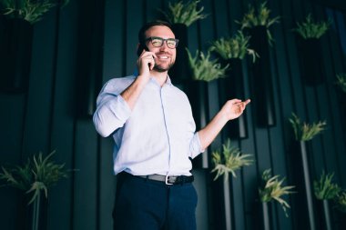 Modern ofisin koridorunda saksı bitkileriyle akıllı telefon konuşmaları yaparken gülümsemenin ve el kol hareketleri yapmanın düşük açısı.