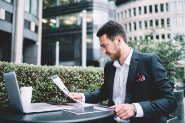 Şehir merkezindeki kafe terasında bilgisayarla çalışırken önemli şirket istatistiklerini kâğıt üzerinde analiz etmekle meşgul düşünceli bir çalışan.