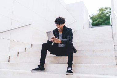Günümüz modası geçmiş esmer, Afro-Amerikalı bir adam gündüz vakti şehir merdivenlerinde otururken tablet üzerinde geziniyor.