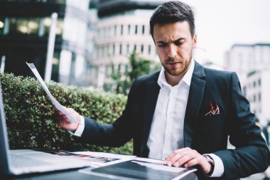 Şehir terasında dizüstü bilgisayarla uğraşırken resmi takım elbiseli, şirket belgelerini dikkatle inceleyen yoğun bir çalışan.