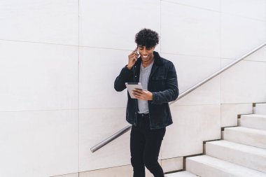 Modern stil güler yüzlü genç esmer Afrikalı Amerikalı adam telefonda konuşuyor ve tablet kullanıyor. Gün ışığında şehir merdivenlerinde dikilirken.