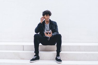 Tabletle merdivenlerde oturmuş kulaklıkla müzik dinlerken beyaz duvara bakan Afro-Amerikalı genç yakışıklı bir adam.