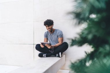 Neşeli, mutlu, yakışıklı, gündelik giyinmiş siyah erkek. Tablet ve kulaklıklarla bacak bacak bacak üstüne atmış ve dışarıdaki kameraya bakıyor.