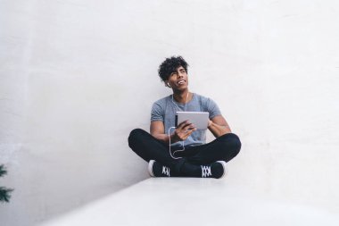 Neşeli, yakışıklı, genç, Afro-Amerikan erkek, gündelik giysiler içinde bacak bacak bacak üstüne atmış, müzik dinliyor, kulaklıklarını takıyor ve dışarı bakıyor.