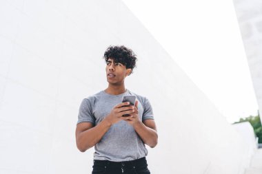 Düşünceli Afro-Amerikalı genç bir erkekten cep telefonuyla sörf yapan ve açık havada bulanık beyaz arka plana bakan.