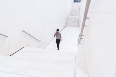 Zeki telefonuyla konuşan, elinde belgelerle beyaz alışveriş merkezinde yürürken gülümseyen, sıradan giyinen neşeli etnik adam.