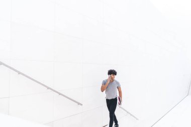 Beyaz iş merkezinde merdivenleri çıkarken akıllı telefondan konuşan neşeli, modern bir etnik adam. Elinde kağıtlar, başka tarafa bakıyor.