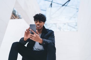 Şık giyinmiş, kıvırcık saçlı, beyaz, modern bir binada otururken akıllı telefon kullanan neşeli, Afro-Amerikan bir adam.