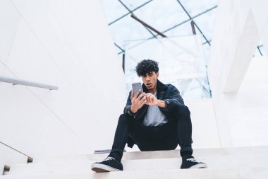 Kafası karışmış, şık giyinmiş, kıvırcık saçlı, beyaz, modern bir binada merdivenlerde otururken akıllı telefon kullanan Afro-Amerikan bir adam.