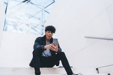 Beyaz modern binada merdivenlerde otururken kıvırcık saçlı, akıllı telefon kullanan şık giyinmiş afro-amerikalı bir adam.