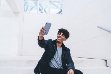 Şık takım elbiseli mutlu kıvırcık siyah adam elinde akıllı telefonuyla büyük beyaz odada oturmuş selfie çekiyordu.