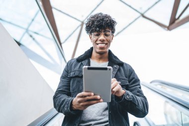 Optik gözlüklü neşeli Latin adam yürüyen merdivende aşağı iniyor ve dokunmatik ped aracılığıyla online sohbetten heyecanlanıyor, milenyum pozitif erkek blogcu mesajlar alıyor