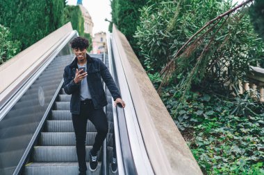 Moda kıyafetli pozitif hipster adam şehir yürüyen merdiveninde aşağı iniyor ve takipçilerle çevrimiçi iletişim kurmak için modern cep telefonu aygıtı kullanıyor, medya dosyalarını paylaşmak için neşeli erkek blogger ağı