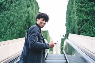 Siyah ceketli ve kot pantolonlu, yeşil ağaçlara bakan yeşil ağaçlara karşı asansörde gezen modaya uygun etnik kıvırcık erkek. 