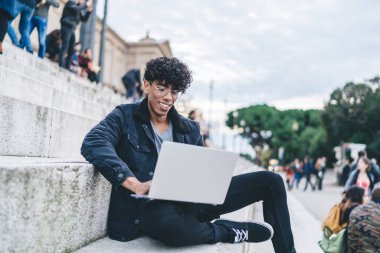 Gözlüklü mutlu etnik adam sıradan kıyafetler giyiyor. Şehir sokaklarında merdivenlerde arka planda insanlarla otururken bilgisayarını faiziyle kullanıyor.