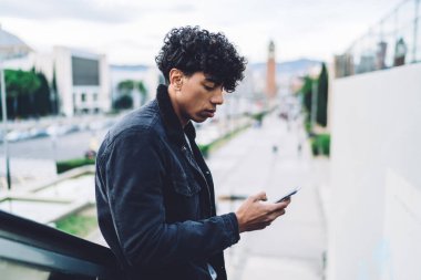 Kıvırcık saçlı, akıllı telefonuyla yürüyen merdivende bulanık arka planda mesajlaşan duyarsız, genç, Afro-Amerikan bir adam.