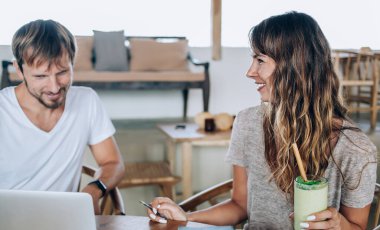 Beyaz tişörtlü genç adam dizüstü bilgisayar kullanıyor ve çekici kadın masada oturup sohbet ederken kokteyl içiyor.