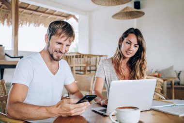 Beyaz tişörtlü neşeli bir adam akıllı telefon kullanıyor ve güzel bir kadın kafede oturmuş kahve içerken dizüstü bilgisayarda geziniyor.