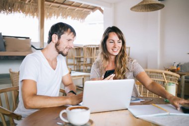 Gülümseyen genç adam ve kadın bir fincan kahve ve dizüstü bilgisayarla masada oturuyor ve akıllı telefon ve arka plandaki sazdan çatılı defterlerle çalışma stratejisi planlıyor.