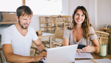 Erkek bilgisayarını karıştırırken ve kadın akıllı telefon kullanırken ve gün ışığında kameraya bakarken, mutlu ve serbest çalışan çift tropikal villada çalışıyor.