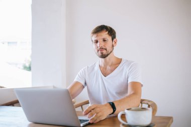 Beyaz tişörtlü, beyaz tişörtlü, beyaz duvarın yanındaki tahta masada oturan ve bilgisayarda yazı yazan orta yaşlı, tıraşsız bir adam.