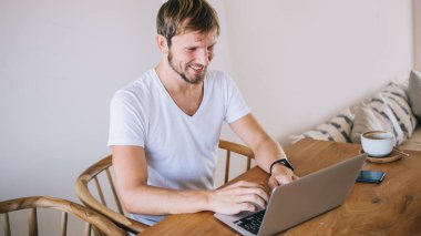 Beyaz tişörtlü, akıllı telefonları olan, dizüstü bilgisayarda sörf yapan ve dairede kahvenin keyfini çıkaran mutlu yakışıklı adamdan.