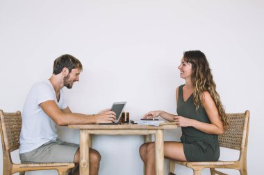 Kafede beyaz arka planda, gülümseyen güzel bir kadının önünde dizüstü bilgisayar kullanan sıradan giysili, yakışıklı bir adamın yan görüntüsü.