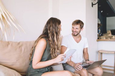 Ellerinde not defteri olan mutlu bir kadın ve mutlu bir erkek dizüstü bilgisayarla kanepede oturmuş birbirlerine bakıyor.