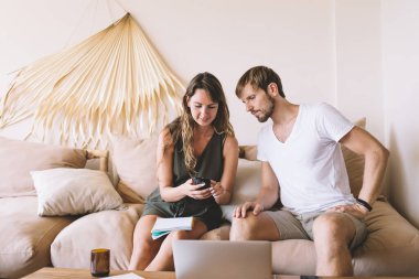 Endişeli yakışıklı erkek, evdeki kanepede oturan çekici bir bayanın ellerinde modern akıllı telefon ekranına dikkatle bakıyor.