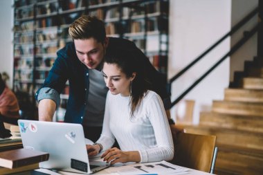 Kot ceketli bir erkek üniversite projesinde çalışan dizüstü bilgisayarı gösteriyor ve kütüphanede masasında oturan bir kadın var.