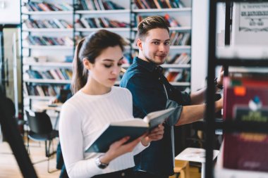 Genç, zeki, konsantre olmuş, günlük giysiler içinde kitap seçen ve okuyan öğrenciler, şehir kütüphanesinde kitaplarla karşı raflarda duruyorlar.