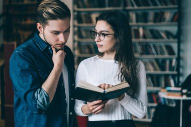Kitaplığın karşısındaki kütüphanede kot gömlekli bir adamla duran beyaz kazaklı genç kadın kitabı işaret ediyor.