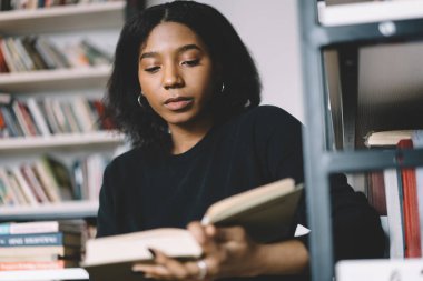 Modern kütüphanede sınava hazırlanmak için günlük kıyafetlerle kitap okuyan Afro-Amerikalı ciddi bir genç kadın.