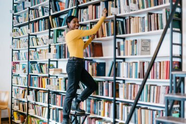 Sıradan giyinmiş, gülümseyen, kameraya bakan, merdivende durup üniversite kütüphanesindeki kitaplıktan kitap alan neşeli siyahi bir kadın.