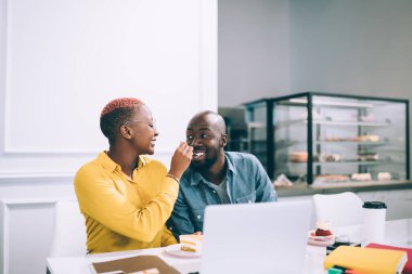 Neşeli, gülümseyen Afro-Amerikan çift kahve dükkanında kahve ve dizüstü bilgisayarla masada oturmuş tatlı ve kek yiyorlar. 