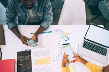 Yukarıdan gelen siyah, meşgul, yüzsüz uzmanlar, ofis masasının etrafında çizimlerle birlikte çalışırken akıllı telefonlara ve boş ekranlı dizüstü bilgisayarlara göz atıyorlar.