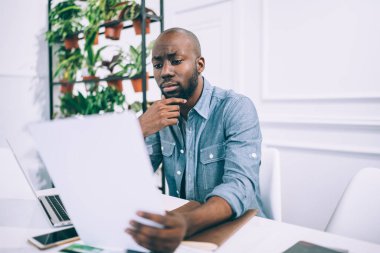 Ciddi bir Afro-Amerikan erkeğine odaklanmış günlük giysiler içinde çene ovuyor belgelerle çalışıyor ve dizüstü bilgisayarla sörf yapıyor yaratıcı stüdyoda masada oturuyor arka planda saksılarda çiçeklerle oturuyor.