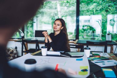 Gündelik giysiler içinde genç bir bayan tasarımcı elinde kağıtlarla masada oturuyor ve tasarım konsepti üzerinde çalışırken meslektaşlarını dikkatle dinliyor. 