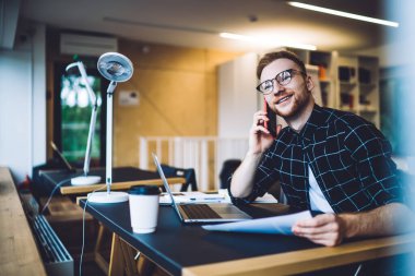 Elinde kağıt olan genç adam bilgisayarla masada otururken gülümsüyor ve düşünüyor ve çağdaş çalışma alanında akıllı telefon konuşmaları yapıyor.