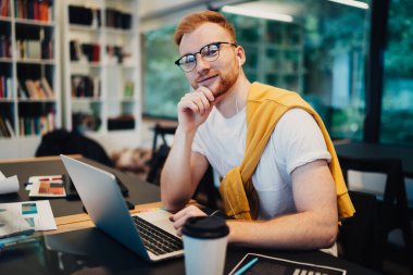 Entelektüel, kızıl saçlı, sıradan beyaz tişörtlü, dizüstü bilgisayarda gezinen arka planda kitaplığı olan bir fincan kahveyle masada oturan bir erkek.