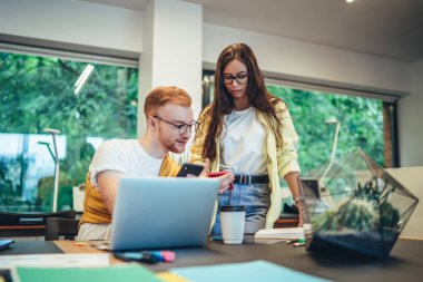 Gülümseyen genç iş arkadaşları gündelik giysiler içinde masada cep telefonları üzerinde ellerinde kahve belgeleri, ofis odasındaki defter akvaryumu ile sörf yapıyorlar.  